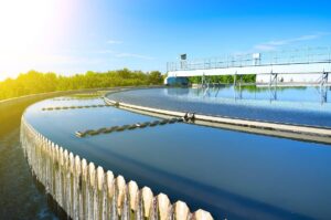 A large, circular water treatment facility in bright sunlight. Clean water flows over a serrated edge into a surrounding gutter. A railing and various equipment are visible on top of the structure,