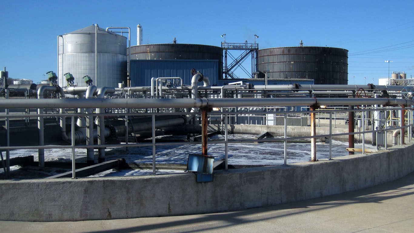 A wastewater treatment plant with cylindrical tanks, metal pipes, and railings hosts a person in protective gear managing the industrial equipment.