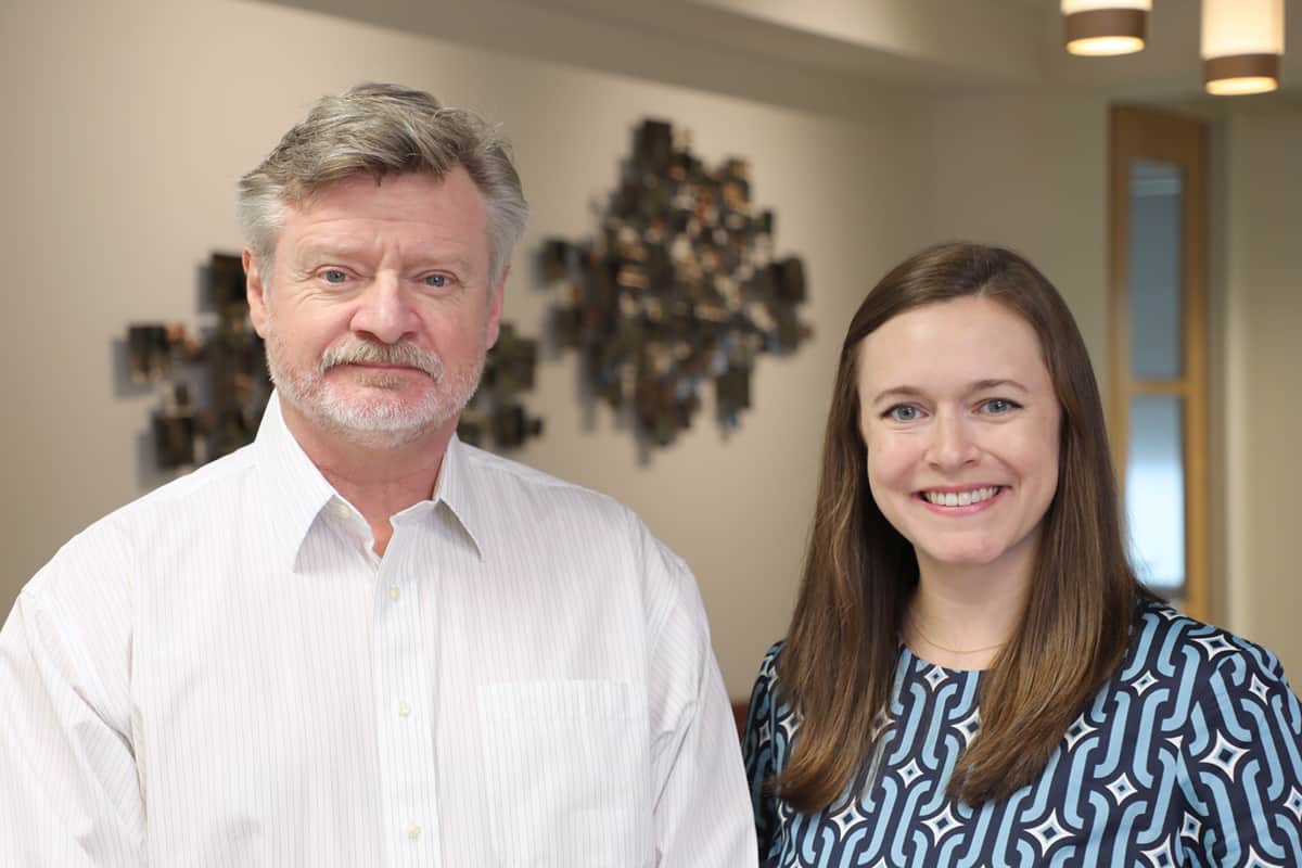 A bearded older man with grey hair and a light-colored button-up shirt stands next to Caitlin, a younger woman with long brown hair, smiling and wearing a patterned blue blouse.