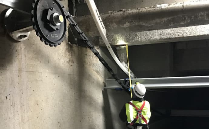 A worker wearing a high-visibility vest and a hard hat is standing in an industrial setting, using a tape measure to gauge a section of a large pipe.