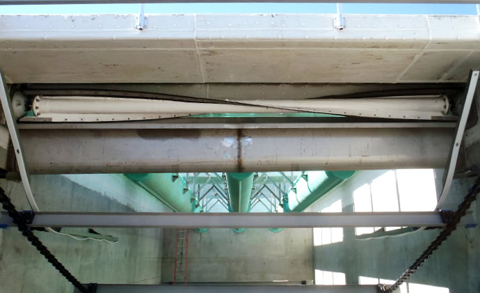 A large industrial pipe carrying water or another fluid is suspended from the ceiling within a concrete structure. Several support beams and green pipes run parallel to the main pipe, and there is a small ladder on the left wall leading upwards.