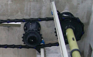 A close-up of a mechanical setup featuring gears, a chain, and rods against a concrete wall. The system includes a chain loop connected to two sprockets and a cylindrical rod.