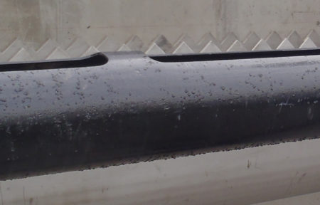 Close-up of a black industrial pipe with water droplets on its surface. The background features a concrete wall with a zigzag pattern.