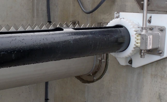 A close-up of an industrial mechanical device showing a large, black cylindrical rod with ridged edges attached to a gear mechanism. The white plastic gear is engaged with the rod.