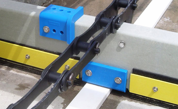 A close-up of a mechanical chain system. A black chain with wheels runs along a set of yellow and white tracks, interfacing with a blue bracket.