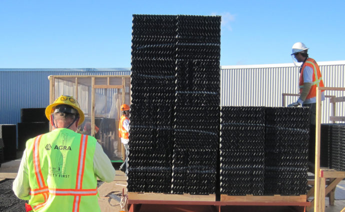 Construction workers in safety gear, including reflective vests and hard hats, are handling large black plastic modular sections at a worksite. One worker stands nearby while another is on a platform beside two tall stacks of the sections.