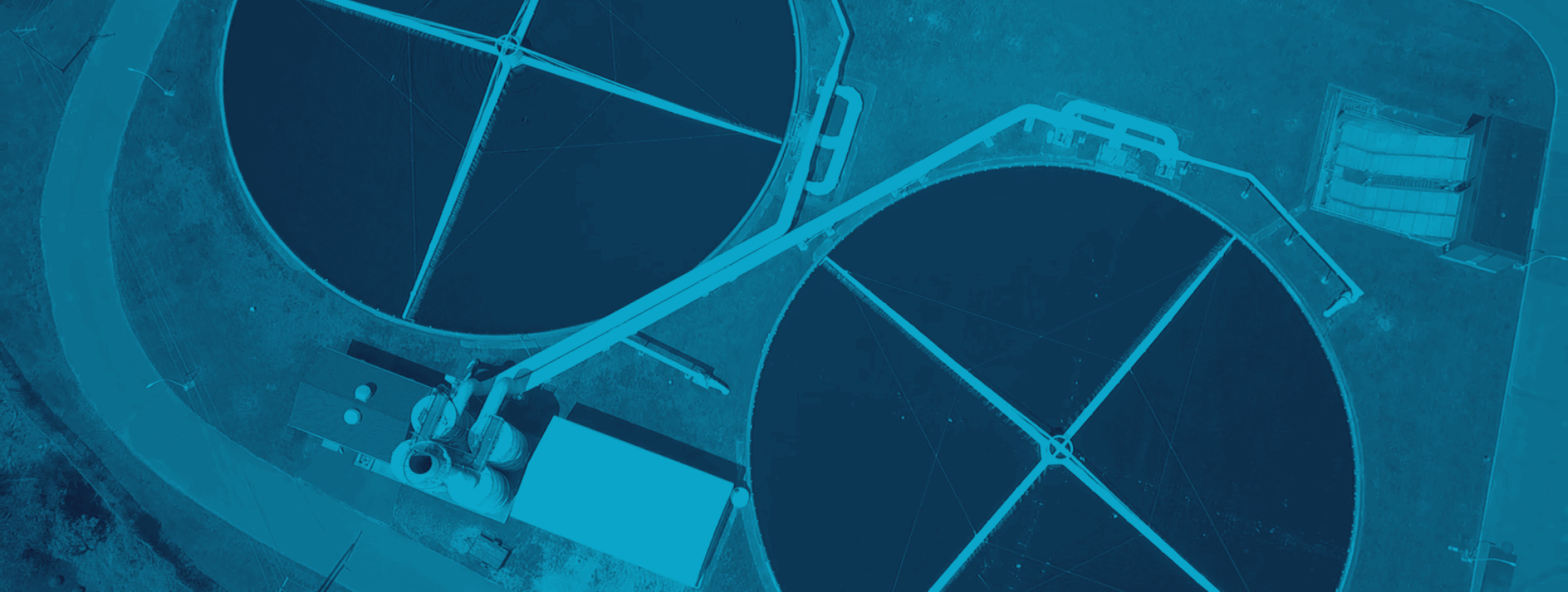 Aerial view of a wastewater treatment facility showing two large circular tanks with crossing structures inside. The image has a blue filter effect.