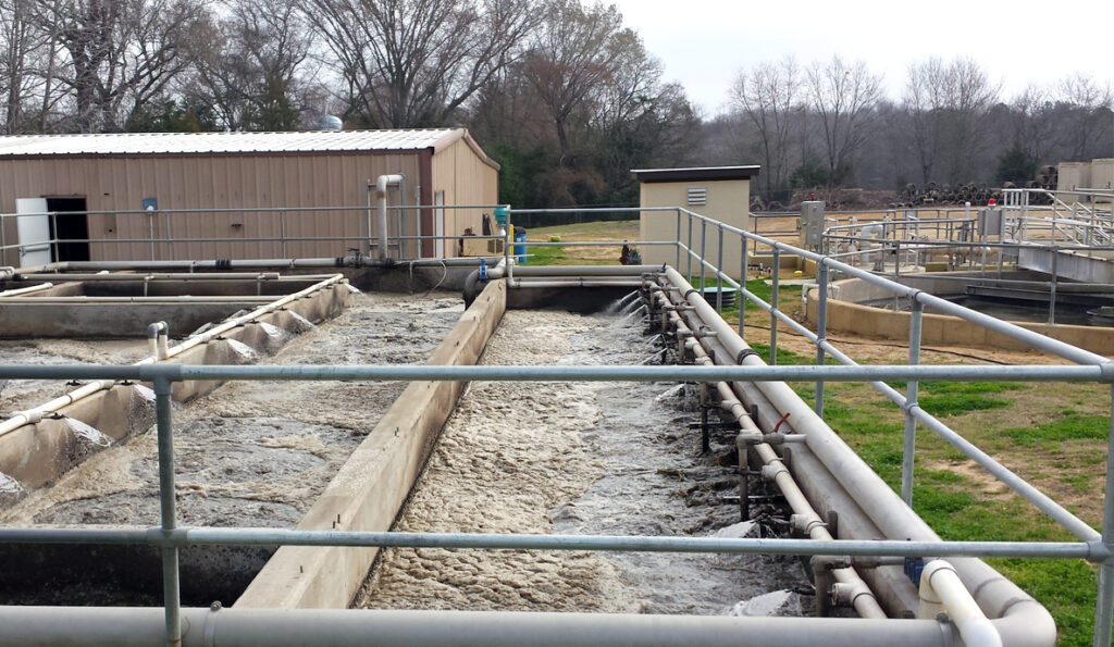 A large outdoor AccuFAS sewage treatment facility with pools of wastewater in concrete basins, separated by metal railings. There are buildings in the background surrounded by trees.