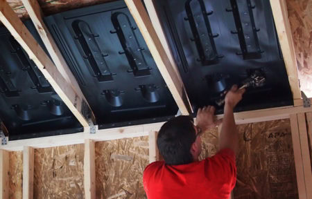 A person in a red shirt installs Accuvent between wooden beams in the ceiling of an unfinished attic, ensuring proper ventilation. The walls have exposed plywood, and the person uses tools to secure the panels in place.