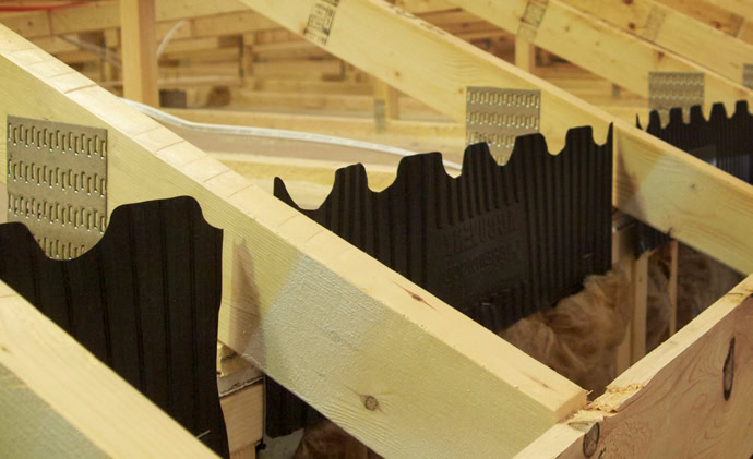 A close-up of an unfinished roof under construction on manufactured homes, featuring wooden trusses and AccuVent black ventilation baffles installed between the rafters. The trusses are fastened with metal connectors, and insulation material is visible in the background.