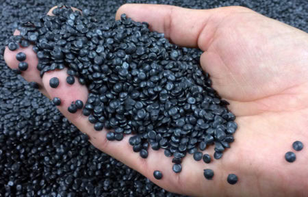 A hand holding a pile of small, black, plastic pellets with more pellets scattered in the background. The round, shiny pellets suggest they are raw materials typically used in plastic manufacturing and recycling processes.