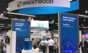 A trade show booth with a large "Brentwood" sign at the top is bustling with activity. Several men are standing and conversing around the booth. Blue panels display text like "We Know Biofilm" and "More Sludge Less Water," promoting SedVac sediment removal technology.