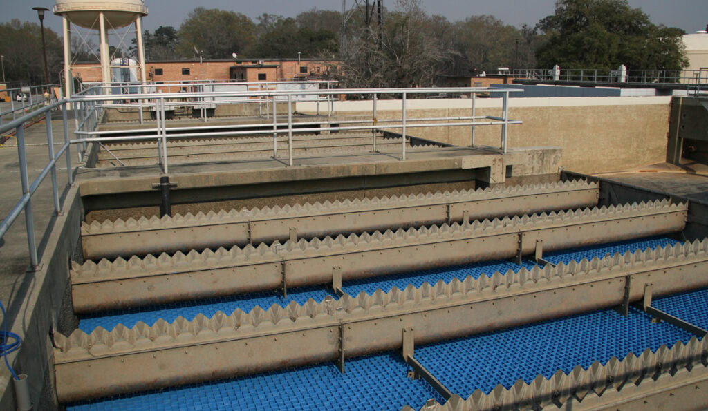 A water treatment facility with a series of rectangular sedimentation tanks. The tanks have jagged, sawtooth edges along the top and blue grids inside.