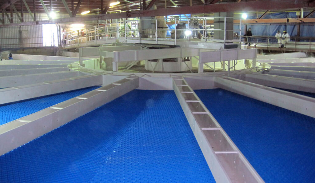 A view of a circular water treatment facility with multiple angled blue filtration surfaces arrayed like spokes from a central point. The facility, featuring the advanced IFR-6000 Series filters, has overhead metal beams and walkways, with people wearing protective suits visible in the background.