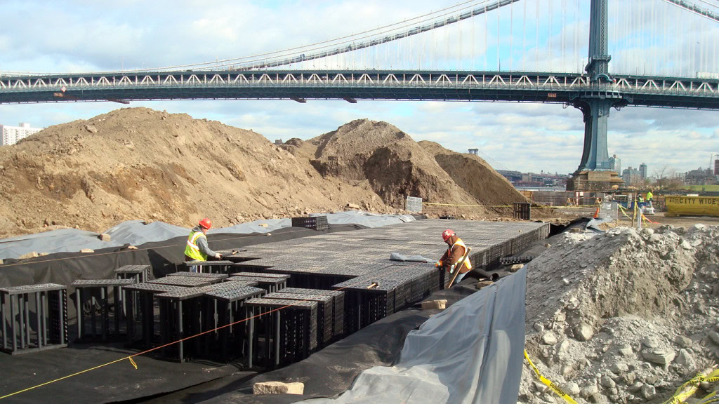 Construction site with workers installing underground structures. Two workers in safety vests and helmets place black plastic components for stormwater management, with mounds of dirt and an overhead bridge in the background. The area is surrounded by protective netting and construction tape.