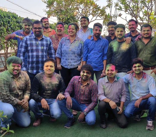 A group of people celebrates Holi, with colorful powder on their faces and clothes. They are smiling and posing for a photo outdoors, surrounded by green trees.