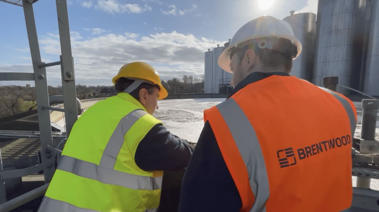 Dos trabajadores con chalecos reflectantes y cascos están de pie en una plataforma industrial. Uno mira hacia otro lado, con casco y chaleco amarillos, mientras que el otro, con chaleco naranja en el que se lee "Brentwood", mira hacia un gran tanque industrial.