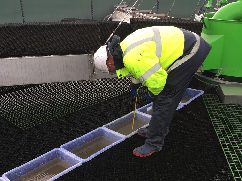 Un trabajador con chaleco de alta visibilidad y casco blanco se inclina sobre una serie de bandejas, utilizando una herramienta para medir o inspeccionar el contenido. Las bandejas están dispuestas sobre un suelo en forma de rejilla y al fondo se ve maquinaria industrial.