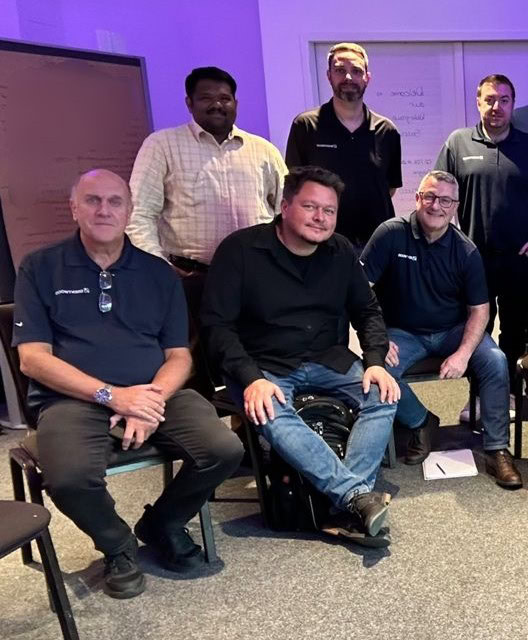 A group of about six men are posing for a photo in an indoor setting with purple lighting. Three are standing at the back while the other three are seated in front.