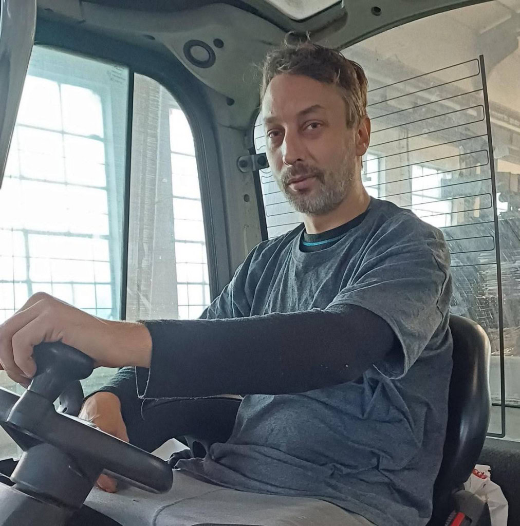 A person with a beard is seated inside a forklift, holding the steering wheel. They are wearing a grey shirt over a long-sleeve black shirt. The background shows large windows and industrial equipment.