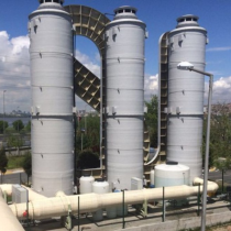 Image of an industrial facility with three tall, cylindrical towers connected by various pipes and structures. The sky is partly cloudy, and the facility is surrounded by some greenery and fencing.