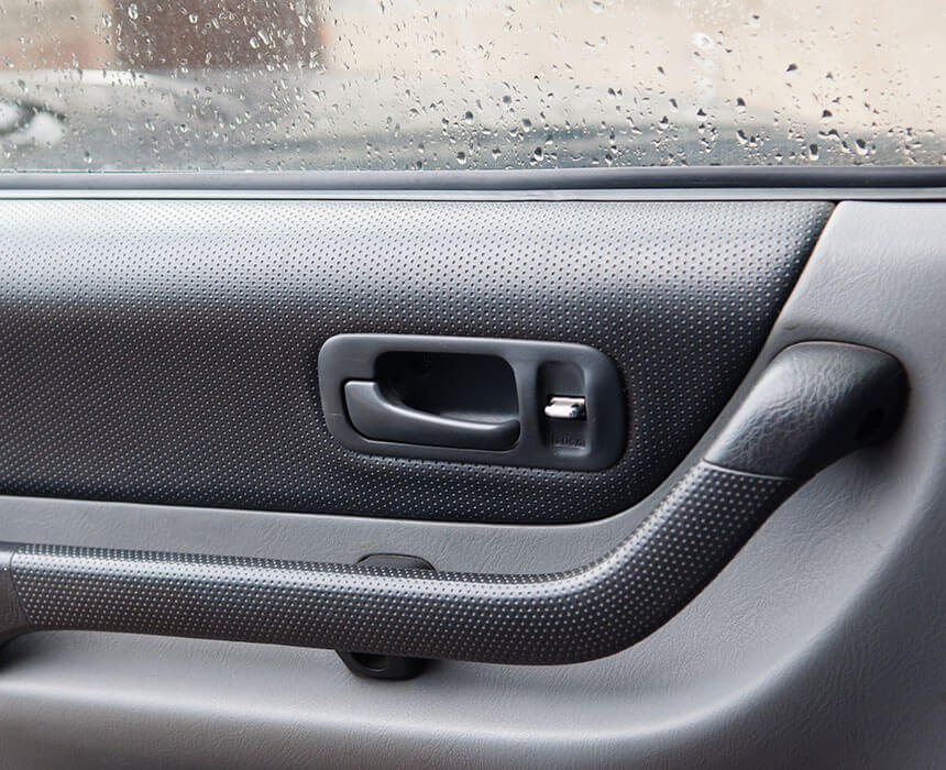 Close-up of a car door interior featuring a door handle and a power window switch, set against a window with water droplets.