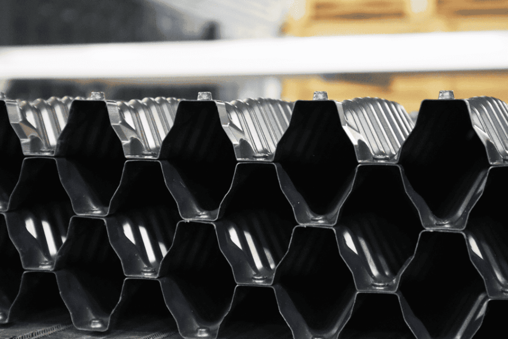 Close-up of multiple stacked, black plastic honeycomb-shaped components, lined with horizontal grooves, creating a repeating hexagonal pattern. The background is blurred, showing part of an industrial setting with wooden pallets.