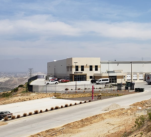 A modern industrial building with loading docks is situated on a hillside with a view of a cityscape in the distance.