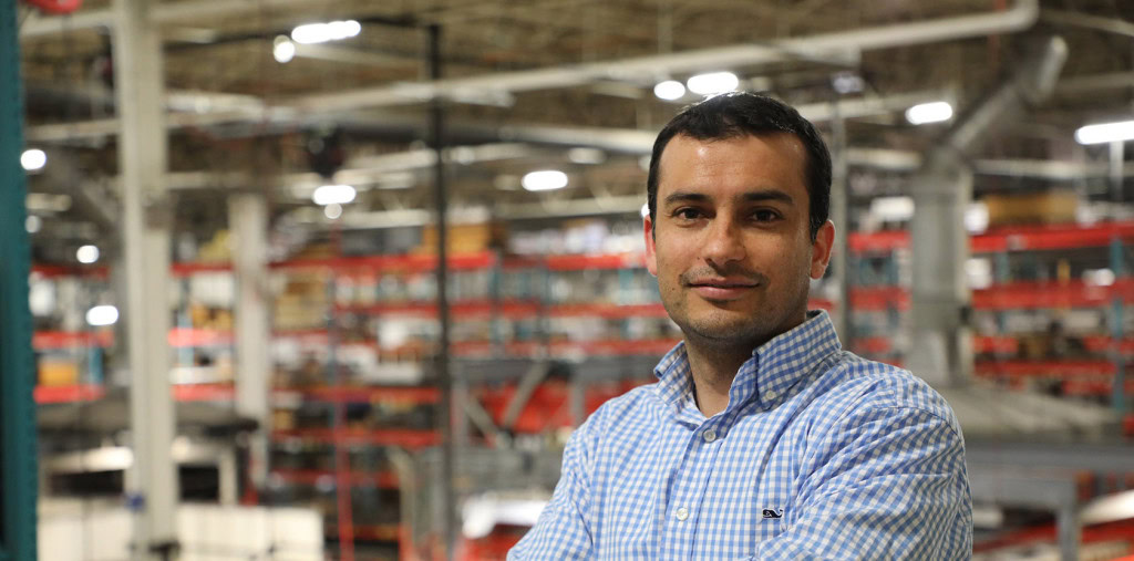 Un hombre con camisa azul a cuadros está de pie con los brazos cruzados, sonriendo ligeramente. El escenario es un espacio interior industrial o un almacén con estanterías y equipos visibles al fondo.