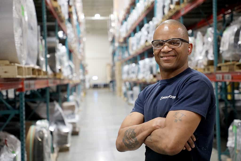 A man wearing safety glasses and a dark T-shirt stands confidently with his arms crossed in a well-organized warehouse aisle.