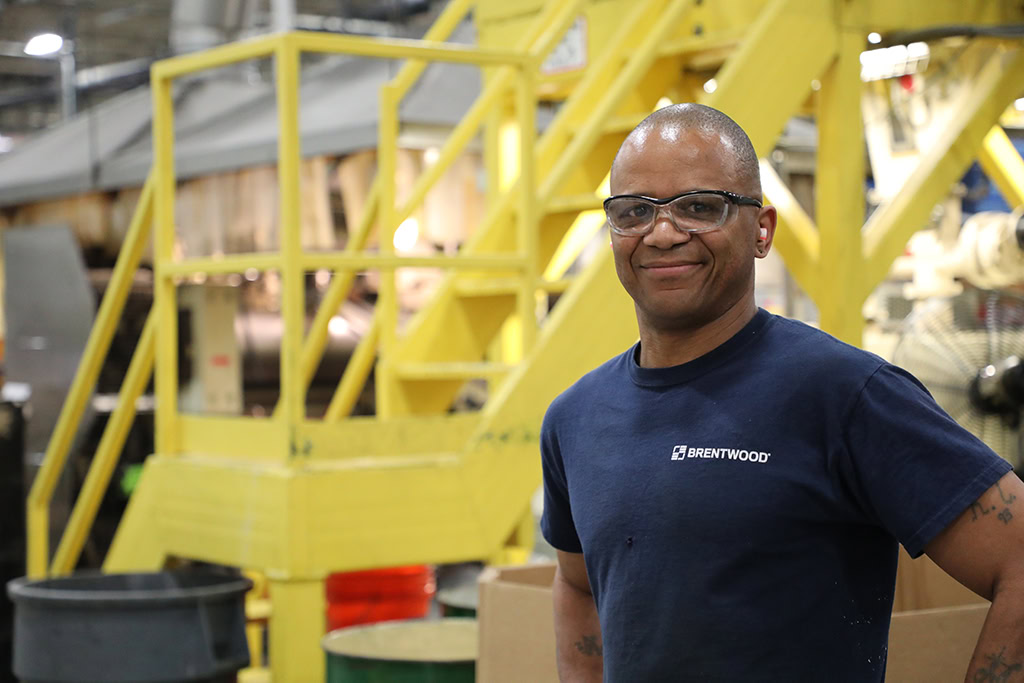 A man wearing safety glasses and a blue "Brentwood" t-shirt stands in an industrial setting. Behind him, there are yellow metal stairs and various machinery.