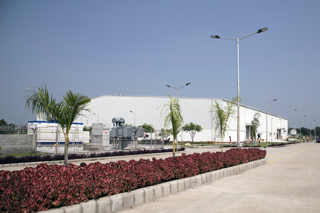 A wide-angle view of an industrial warehouse with a white exterior is showcased. Around the area, you'll find well-maintained landscaping featuring red shrubs, young palm trees, and neatly paved pathways with streetlights.