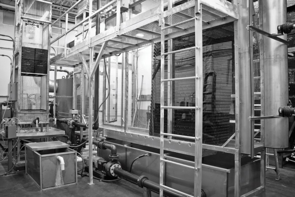 A black-and-white image of an industrial facility featuring large metal machinery, pipes, and a platform with metal railings.