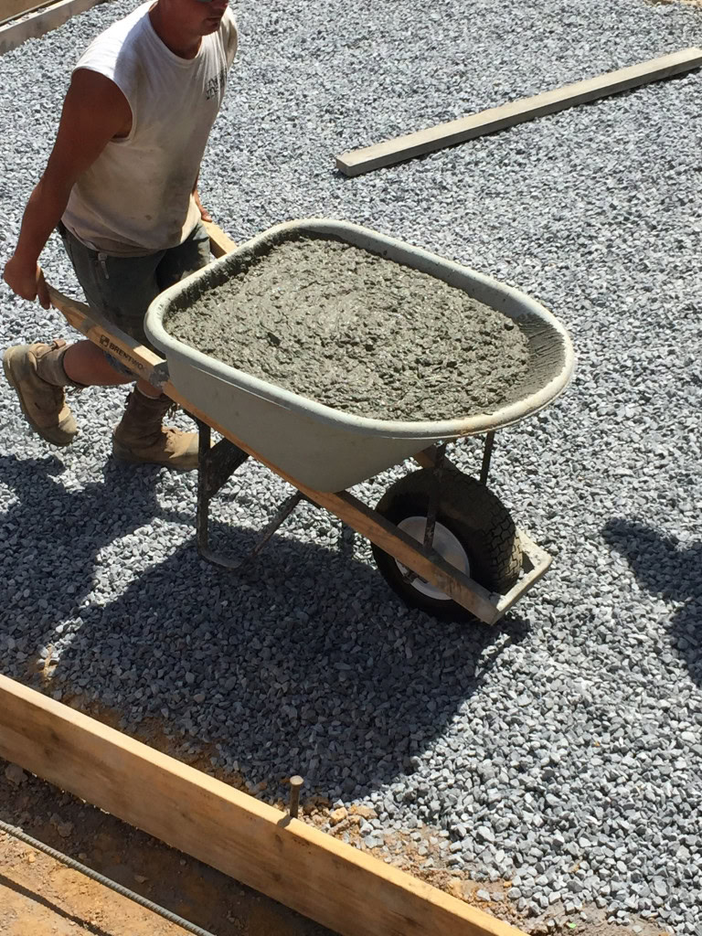 Una persona empuja una carretilla llena de hormigón húmedo sobre un lecho de grava en una obra. Lleva camisa sin mangas, pantalones cortos y botas de trabajo. Al fondo se ven tablones de madera y otros equipos de jardinería.