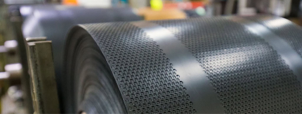 Close-up of perforated metal sheets being rolled up in a factory setting. The metal sheet displays a pattern of evenly spaced small circular holes.