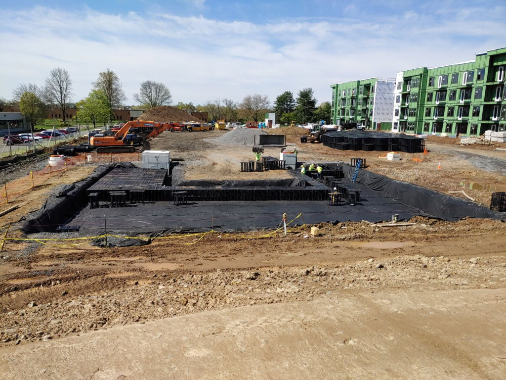 A housing construction site with partially built foundations is surrounded by black tarp. Nearby are construction equipment and materials, including excavators and gravel piles. In the background, partially constructed apartments with green exterior walls are visible.