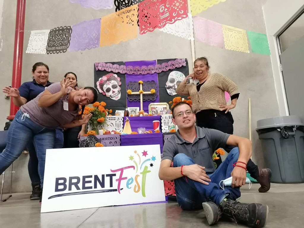 A group of five people pose in front of a colorful Day of the Dead altar decorated with marigolds, skulls, and other traditional items. The altar is set against a gray wall with colorful papel picado hanging above. There is a sign that shows BrentFest