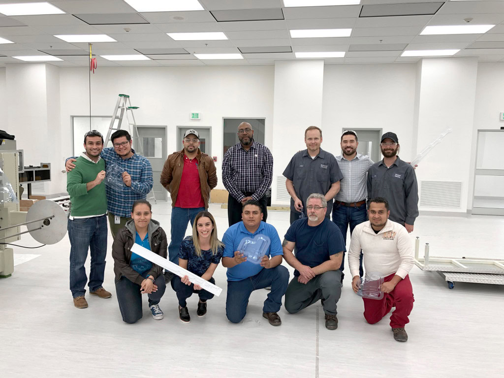 A group of twelve people, including men and women, stand and kneel in a bright, white room, posing for a team photo.