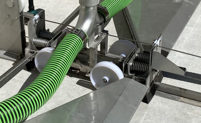 Close-up of a SedVac sediment dredge system with a large green ribbed hose connected to a metallic structure. White plastic wheels and various metal springs are visible as part of the assembly.