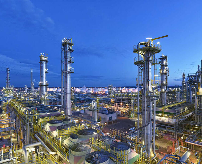 A sprawling industrial refinery complex is shown at dusk, with tall towers, numerous pipes, and various structures illuminated against a deep blue sky.