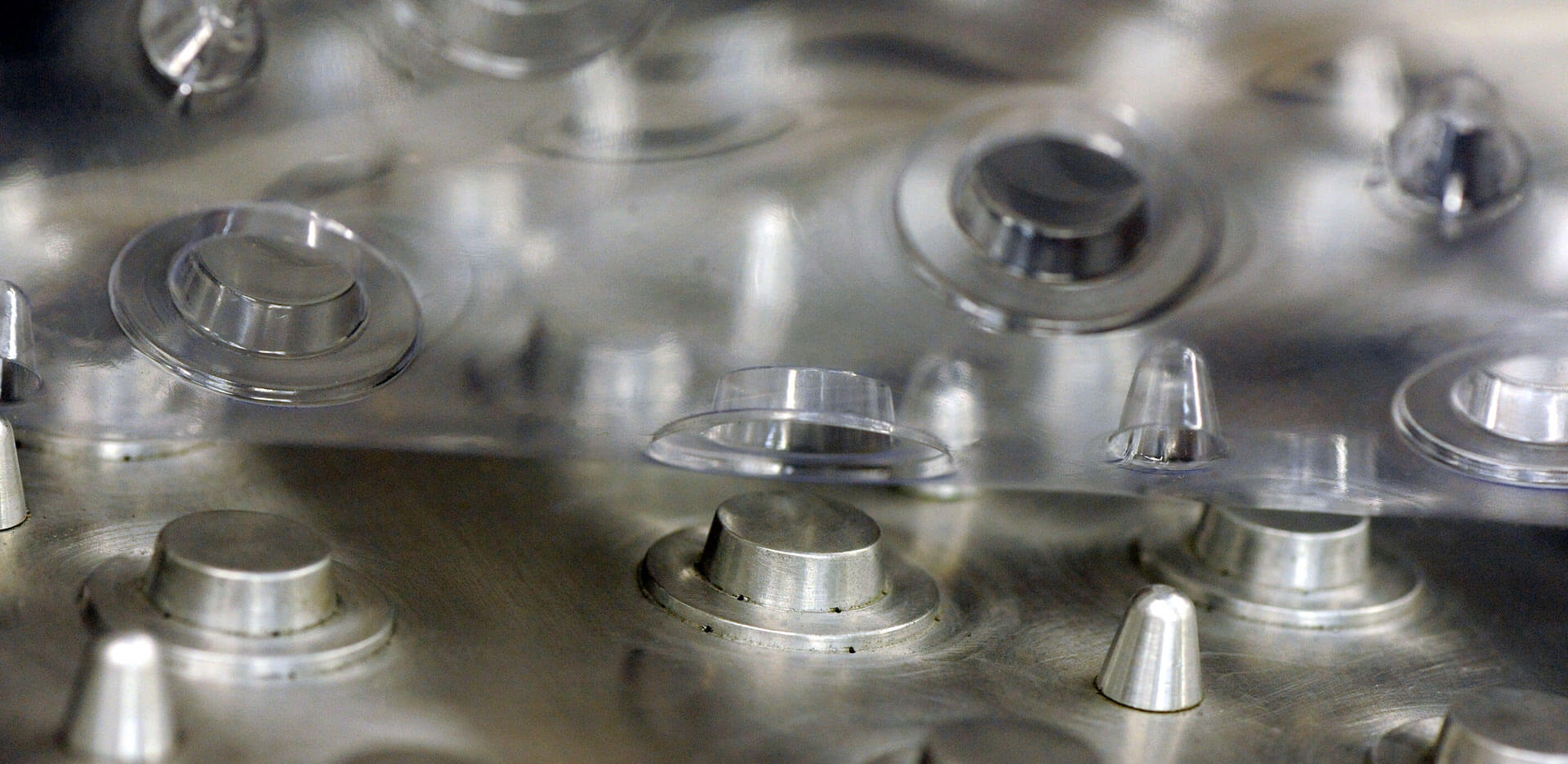 Close-up image of a manufacturing process showing clear plastic blisters being formed on a metal mold. The transparent material, reveals multiple blister cavities with corresponding ridges on the metallic surface below.