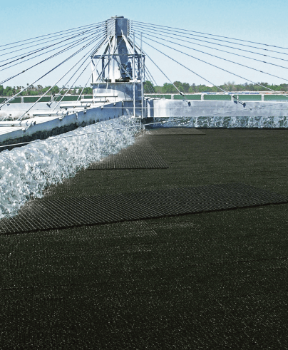 A round, industrial water treatment tank with a structural framework and cables extending outward is shown under a clear blue sky. Water cascades over dark filtration material within the tank.