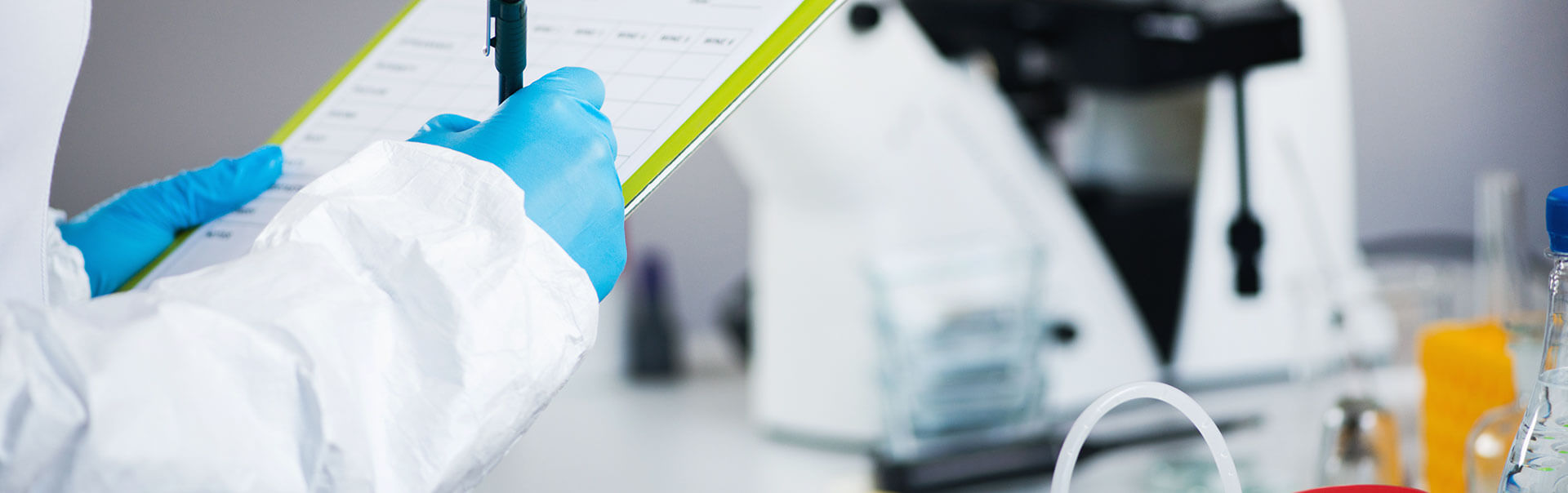 A person wearing a white lab coat and blue gloves writes on a clipboard in a laboratory,