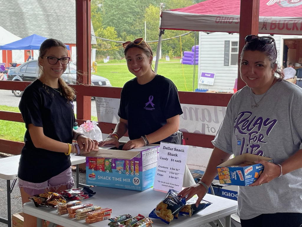 Three smiling individuals standing behind a snack booth. The table is filled with various snacks including chips, candy, and snack bars. A sign on the table lists prices for the snacks. The background shows a tent and other event activities.