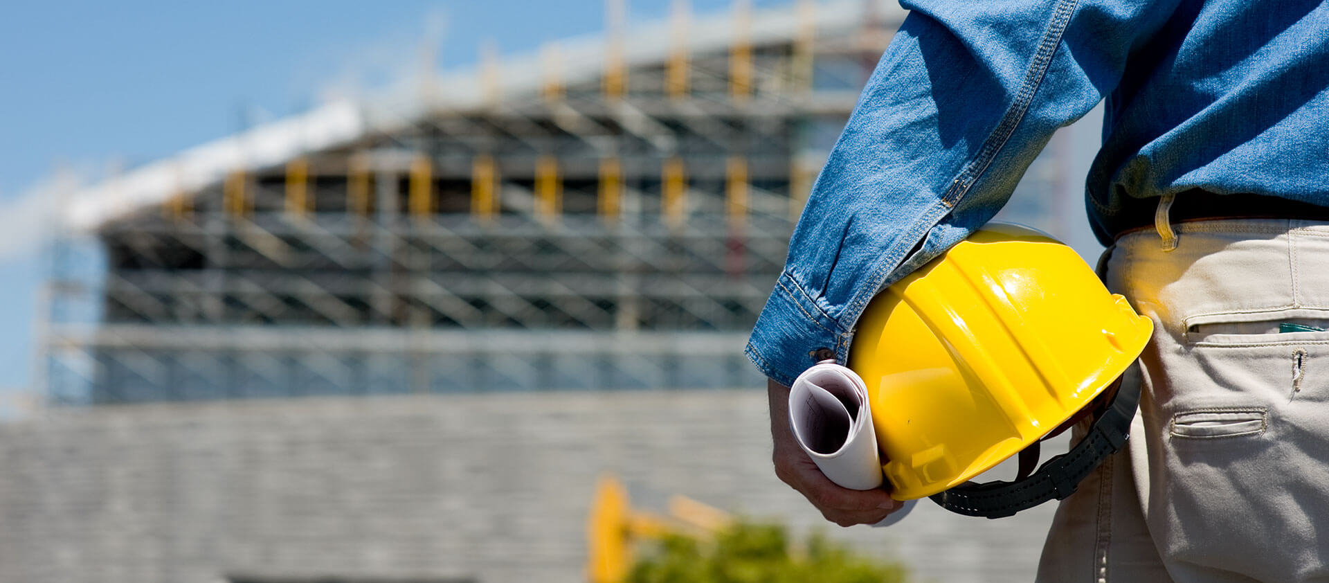 A construction worker stands with their back to the camera, wearing a blue shirt and holding a yellow hard hat and rolled-up blueprints. In the background, there is a large construction site with scaffolding in place.