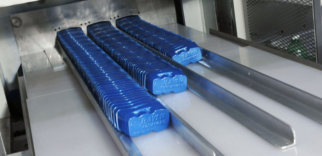 Close-up of a packaging machine focusing on blue rectangular items stamped with "BAYER Advanced" aligned in parallel rows on a conveyor belt.