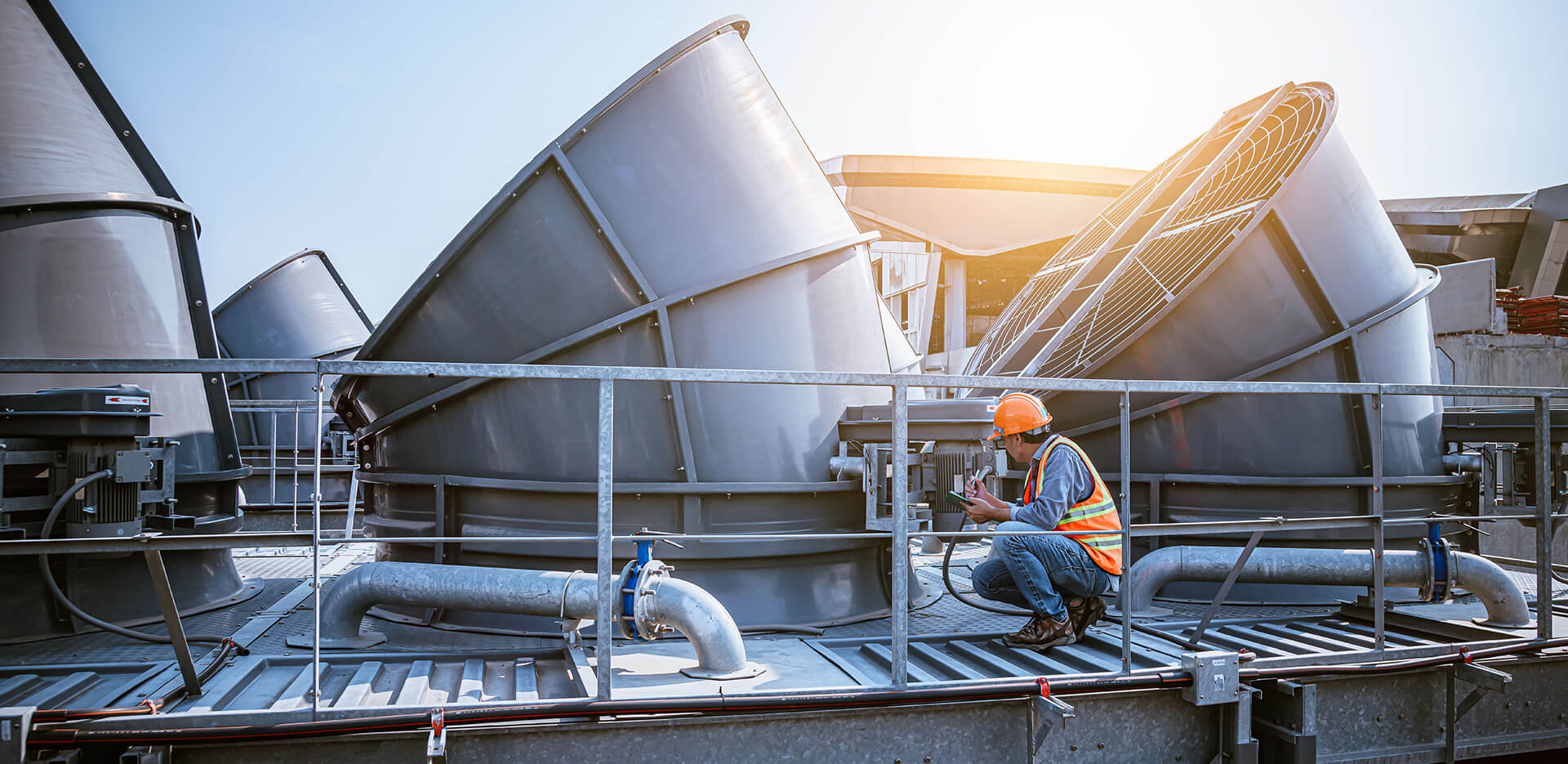 A worker wearing a hard hat and a high-visibility vest uses tools while inspecting or maintaining large industrial cooling units on a rooftop under sunlight.