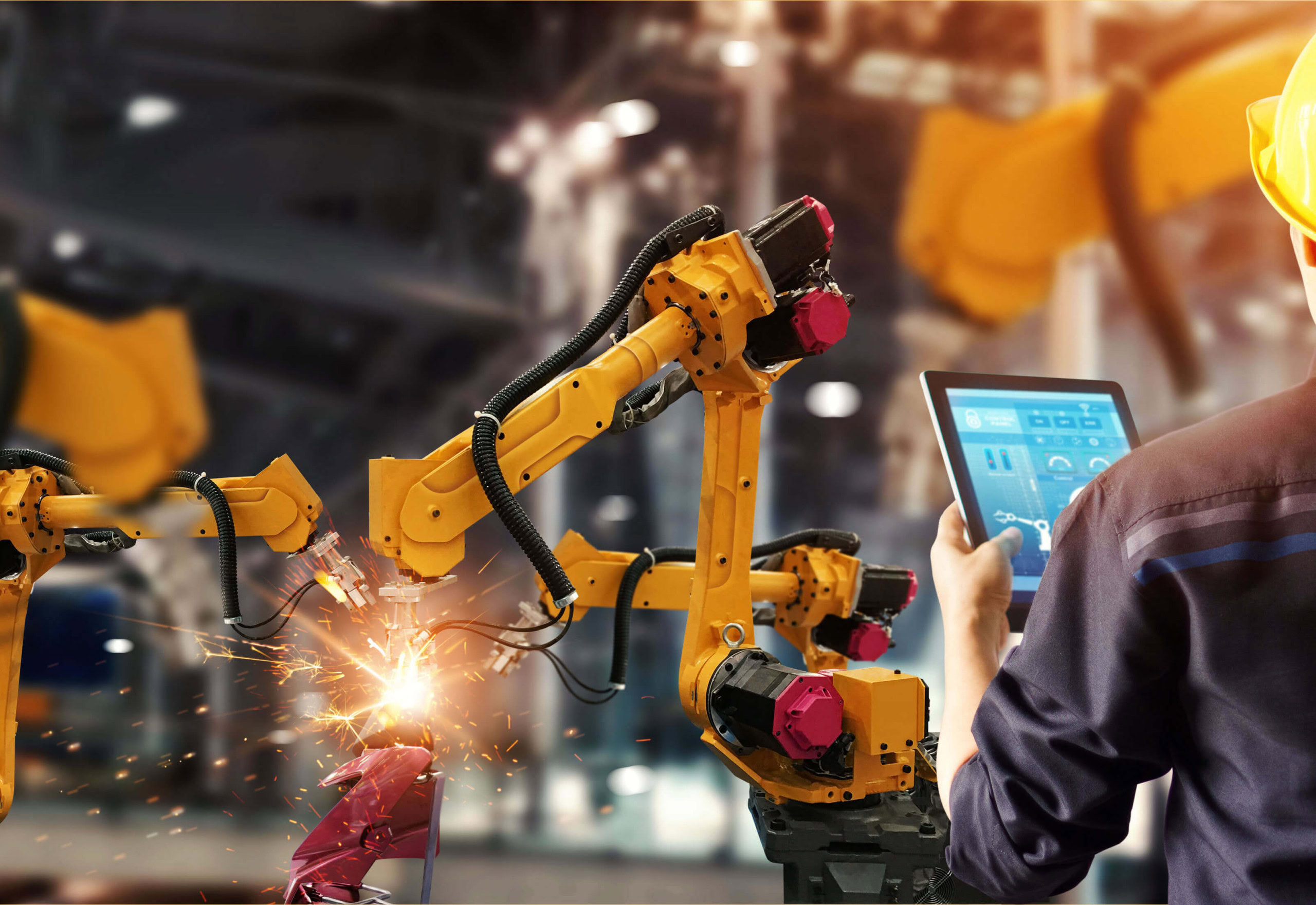 A person in a yellow hard hat operates a tablet while robotic arms perform welding tasks in an industrial setting.