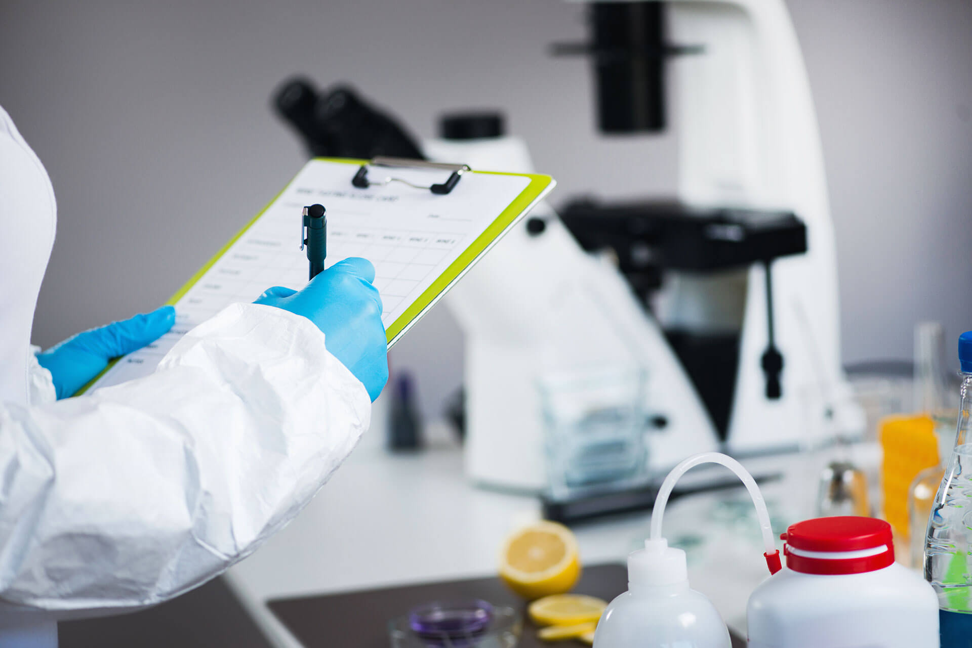 A person wearing blue gloves and a white lab coat holds a clipboard and pen, making notes in a laboratory. A microscope and various lab equipment are visible in the background.