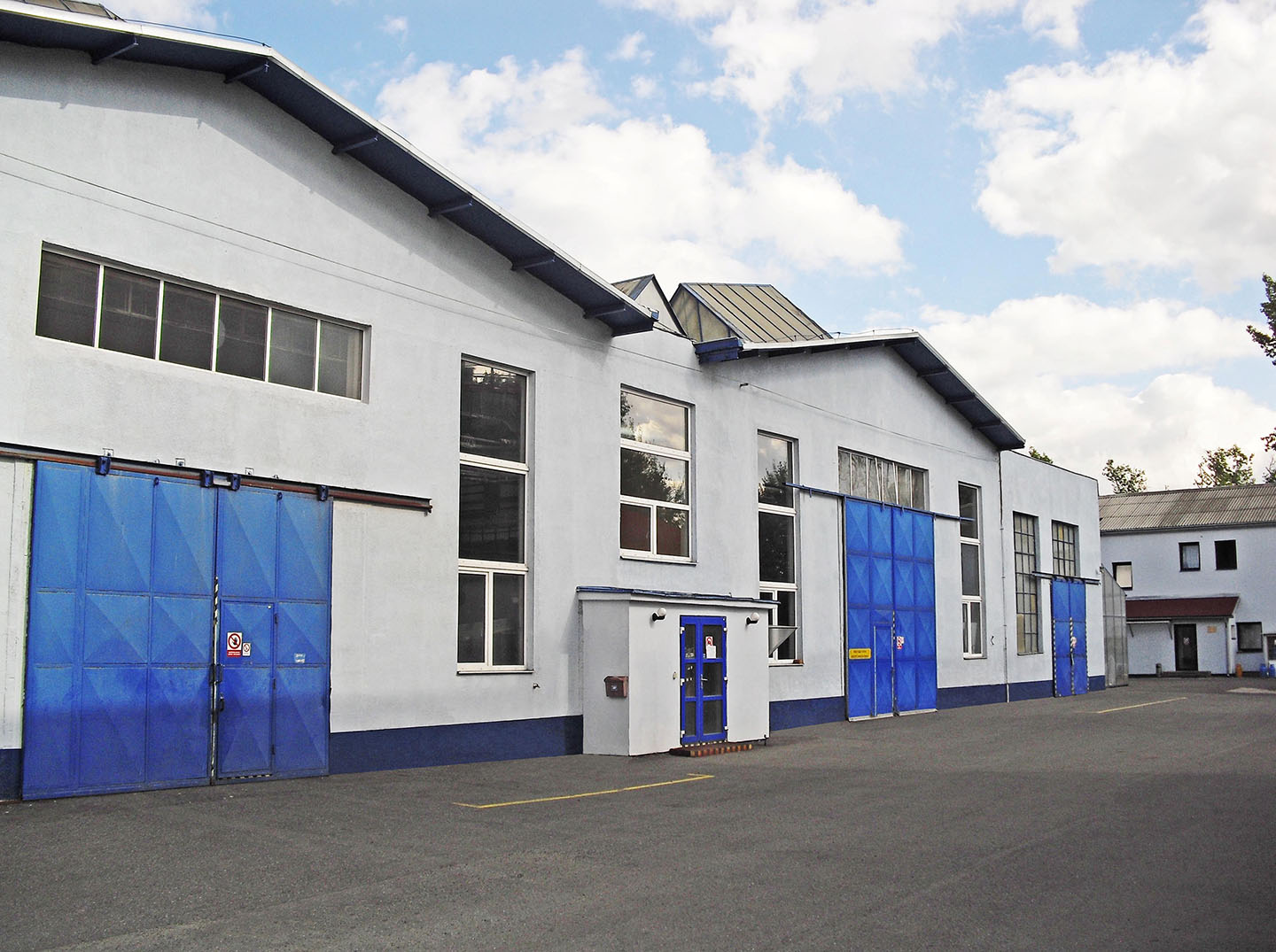 Un edificio industrial con una fachada blanca y grandes puertas azules, incluyendo estilos enrollables y abatibles. Varias ventanas bordean las partes superiores del edificio.